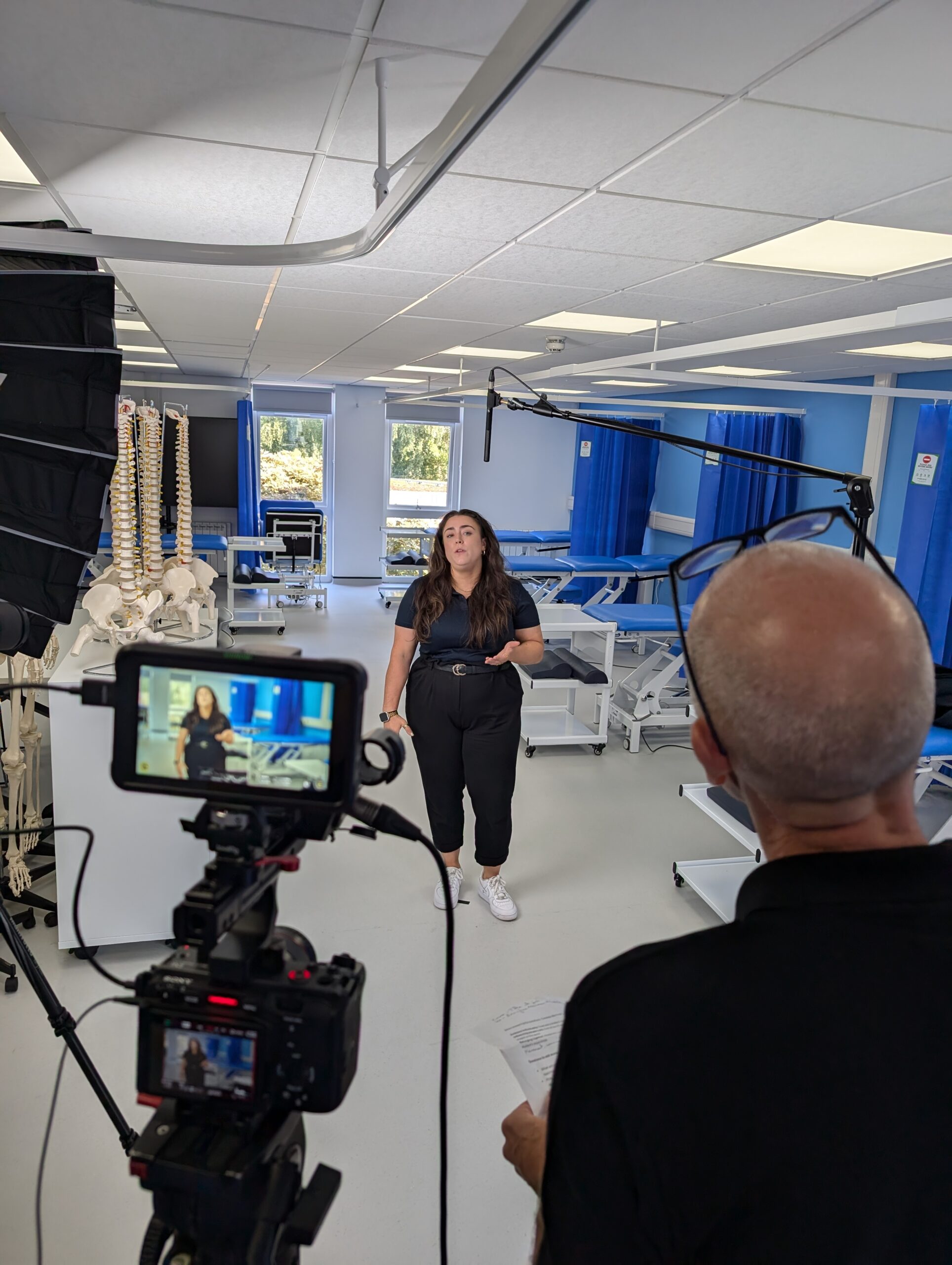 Behind-the-scenes of a promotional video shoot for Newman University's Sports Therapy course, featuring a professional setup with lighting, cameras, and a presenter in a clinical classroom setting.